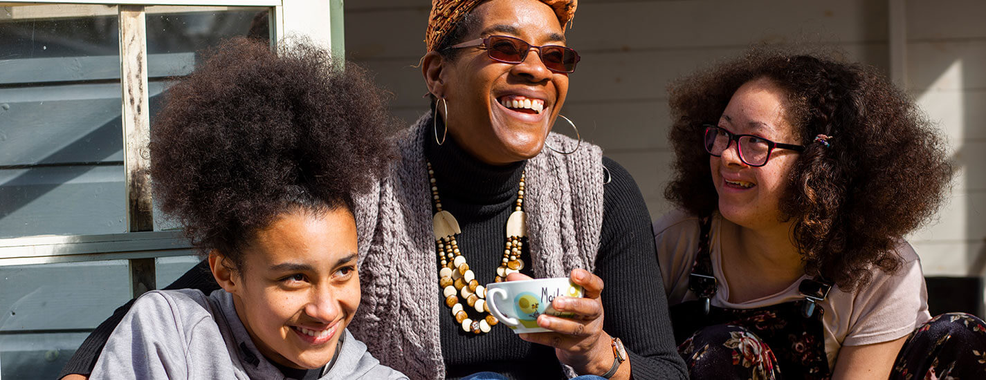 A mom and her two daughters are sitting on their back steps, enjoying coffee and smiling.